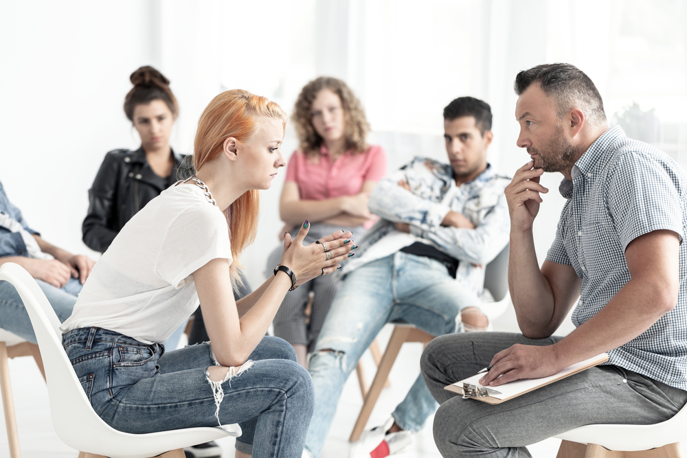A fiery-haired young woman with a rebellious spirit engages in conversation with a counselor during a group therapy session at a rehabilitation center.