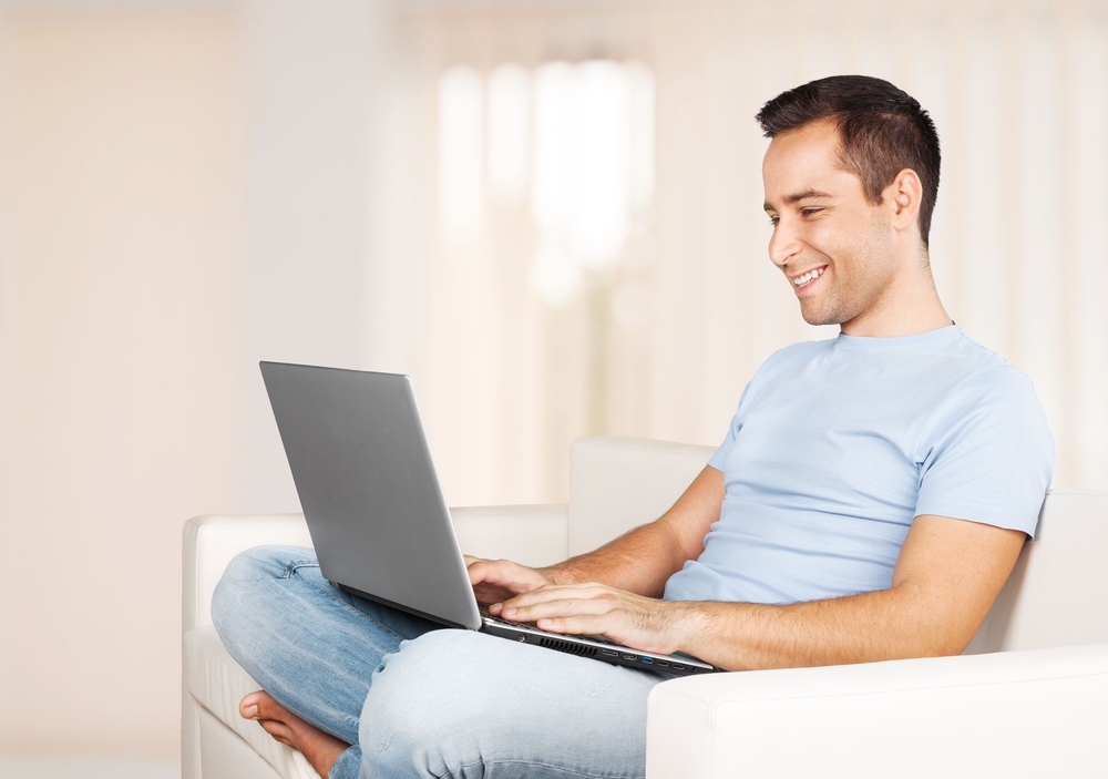 The smiling, sober man is sitting on the sofa and looking at the laptop after receiving mental health treatment.