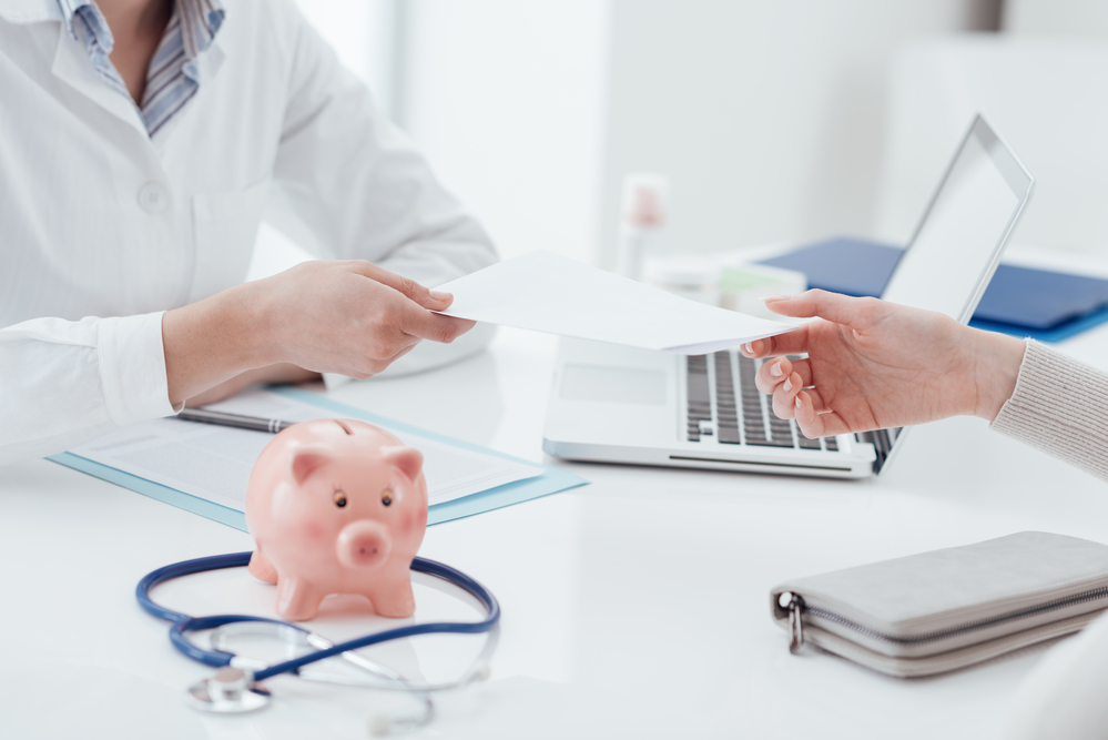 Female doctor working at office desk with stethoscope and piggy bank on the foreground: health insurance concept
