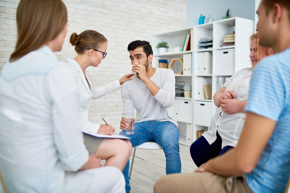 Portrait of female psychiatrist comforting crying young man in group rehab therapy session with other patients sitting in circle around them